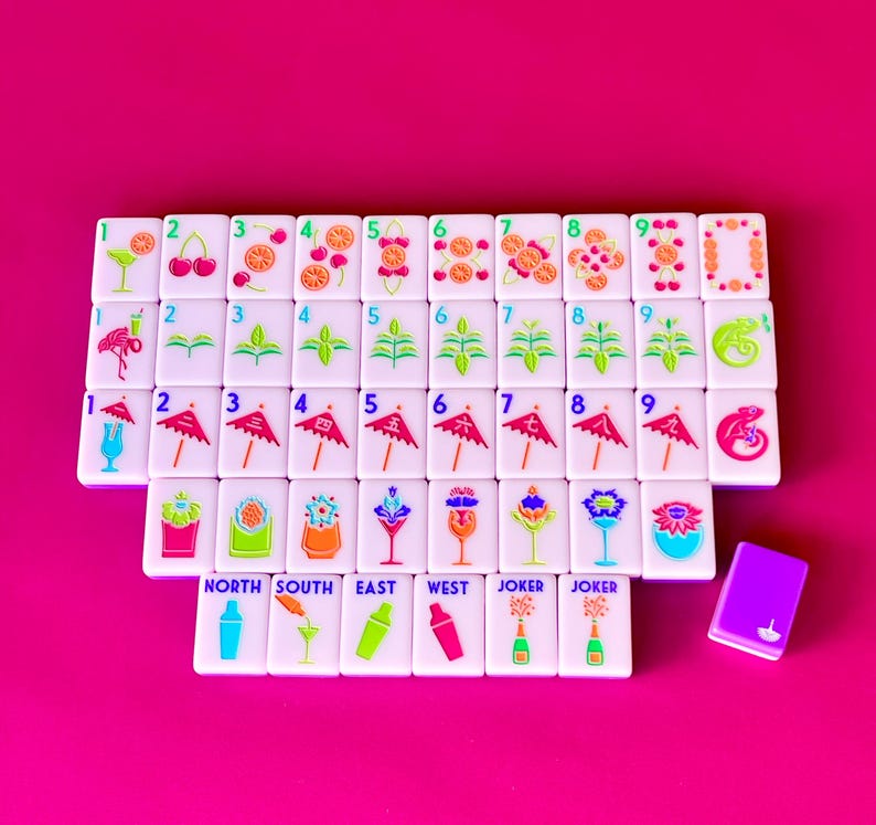 May include: A set of white Mahjong tiles with colorful illustrations of cocktails, fruit, and plants. The tiles are arranged on a bright pink surface, with a small purple tile box. The text "NORTH SOUTH EAST WEST JOKER JOKER" is visible.