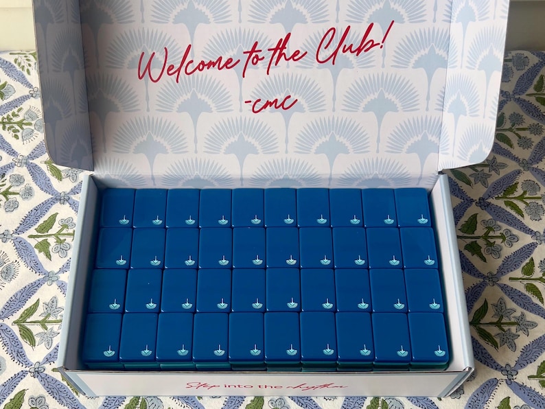 May include: An open box filled with blue rectangular tiles, each with a small white design. The box interior has a light blue pattern and the words "Welcome to the Club!" in red. The box is on a patterned surface.
