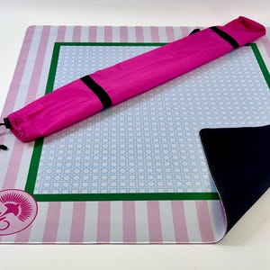 May include: A pink and white striped yoga mat with a light blue and white patterned center. A bright pink carrying case with black straps rests on the mat. The mat has a green border and a logo in the corner.