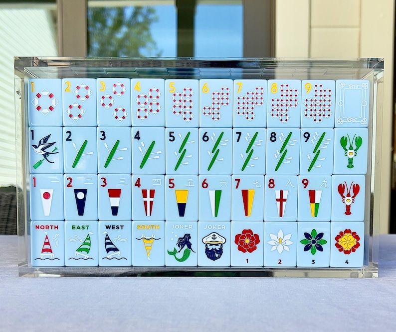 May include: A set of light blue Mahjong tiles with various symbols and numbers, including nautical images like sailboats and compass points. The tiles are displayed in a clear acrylic case.
