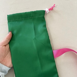 Puede incluir: Una bolsa verde con cord&oacute;n y correa rosa. La bolsa est&aacute; hecha de un material liso y brillante. El cord&oacute;n es blanco con un cord&oacute;n rosa.