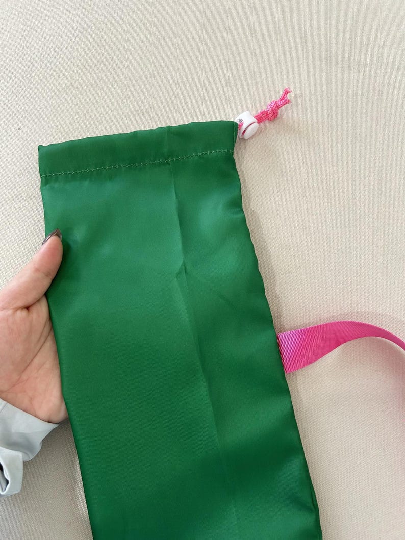 Puede incluir: Una bolsa verde con cord&oacute;n y correa rosa. La bolsa est&aacute; hecha de un material liso y brillante. El cord&oacute;n es blanco con un cord&oacute;n rosa.