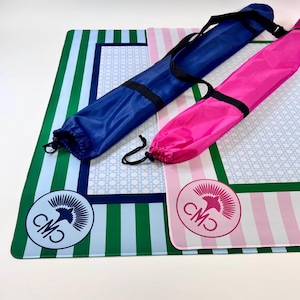 May include: Two colorful beach mats with matching carrying cases. One mat features green and blue stripes, the other pink and white stripes. Both have a circular logo and a blue or pink carrying case.