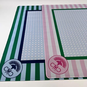 May include: Two rectangular placemats. One has green and blue vertical stripes, a light blue center with a white lattice pattern, and a circular logo. The other has pink and white stripes, a light blue center with a white lattice pattern, and a circular logo.