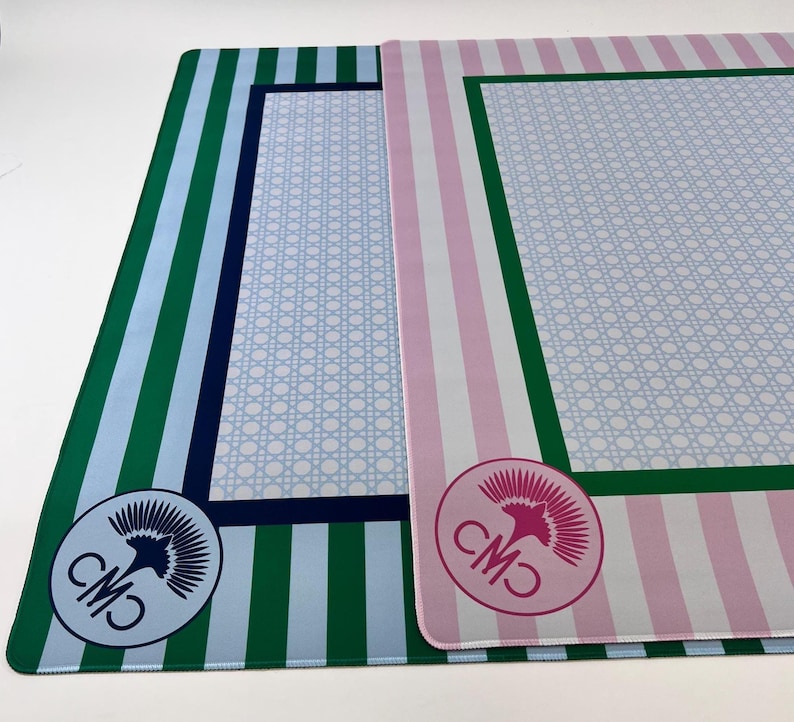 May include: Two rectangular placemats. One has green and blue vertical stripes, a light blue center with a white lattice pattern, and a circular logo. The other has pink and white stripes, a light blue center with a white lattice pattern, and a circular logo.