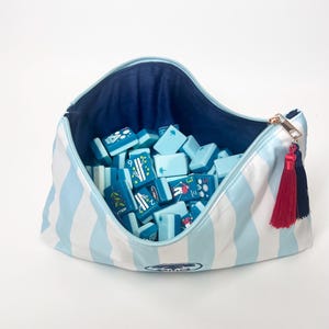 May include: A blue and white striped zippered bag filled with light blue game tiles. The bag has a navy blue interior and a gold zipper pull with red and navy tassels. The tiles have various illustrations.