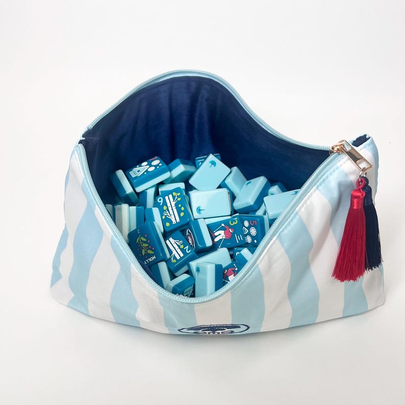 May include: A blue and white striped zippered bag filled with light blue game tiles. The bag has a navy blue interior and a gold zipper pull with red and navy tassels. The tiles have various illustrations.