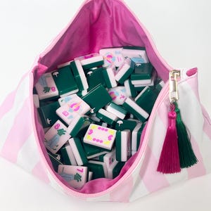 May include: A pink and white striped zippered pouch filled with green and white Mahjong tiles. The pouch has a pink interior and a gold zipper with pink and green tassel accents. The tiles are various colors and feature symbols.