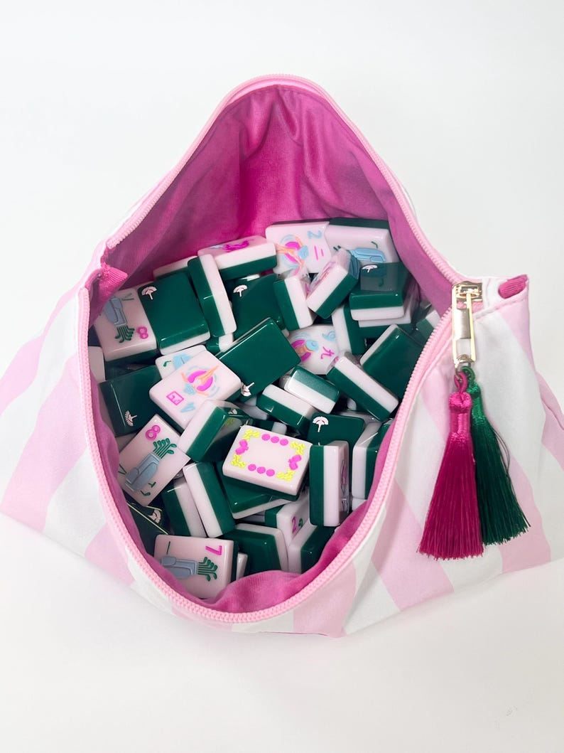 May include: A pink and white striped zippered pouch filled with green and white Mahjong tiles. The pouch has a pink interior and a gold zipper with pink and green tassel accents. The tiles are various colors and feature symbols.