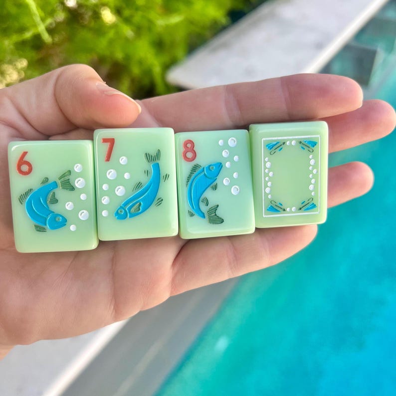 May include: Four light green Mahjong tiles with blue fish illustrations and white bubbles. The tiles are numbered 6, 7, and 8, with the fourth tile featuring a decorative border. The tiles are held in a hand.