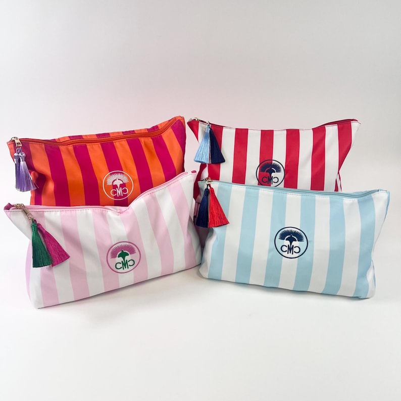May include: Four striped cosmetic bags in shades of pink, orange, red, and blue. Each bag features vertical stripes, a tassel zipper pull, and a circular logo. The bags are arranged on a white surface.