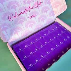 May include: An open box filled with numerous rectangular, purple objects. Each object has a small, white design. The inside of the box lid features a repeating pink fan pattern and the text "Welcome to the Club!" in pink script.