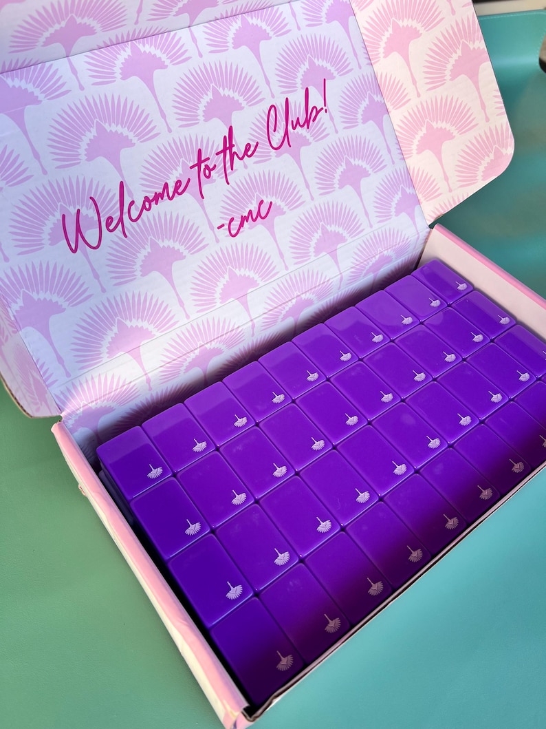 May include: An open box filled with numerous rectangular, purple objects. Each object has a small, white design. The inside of the box lid features a repeating pink fan pattern and the text "Welcome to the Club!" in pink script.