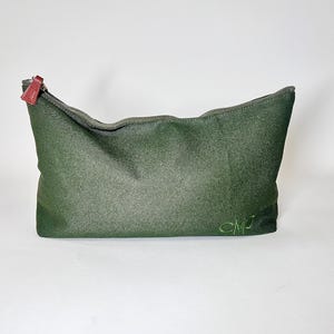 May include: A dark green zippered pouch with a textured surface. The zipper pull is a reddish-brown color. The initials "CMJ" are embroidered in green on the lower right side of the pouch. The pouch is likely used for cosmetics or personal items.