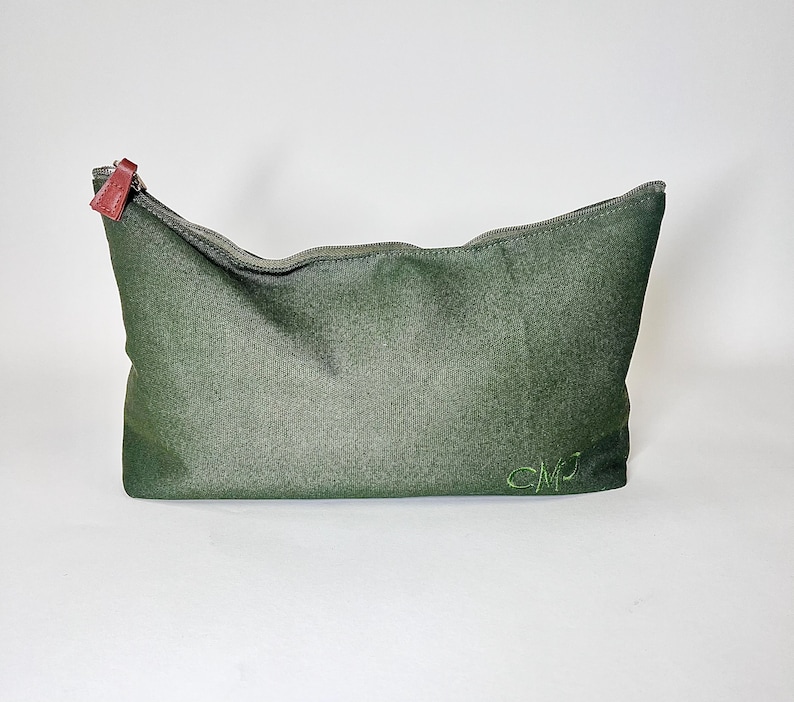 May include: A dark green zippered pouch with a textured surface. The zipper pull is a reddish-brown color. The initials "CMJ" are embroidered in green on the lower right side of the pouch. The pouch is likely used for cosmetics or personal items.