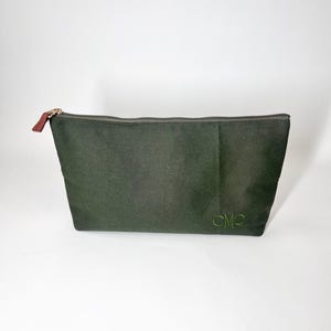 May include: An olive green zippered pouch with a gold zipper and a brown leather pull tab. The pouch has the embroidered initials "CMP" in green on the lower right corner. The pouch is made of a textured fabric.
