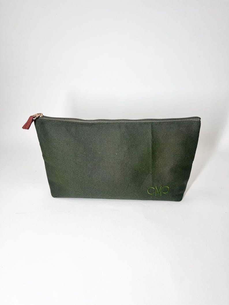 May include: An olive green zippered pouch with a gold zipper and a brown leather pull tab. The pouch has the embroidered initials "CMP" in green on the lower right corner. The pouch is made of a textured fabric.