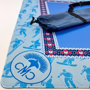 May include: A blue ski-themed mat with a matching carrying bag. The mat features skiers and snowboarders in light blue on a blue background. A decorative border with red, white, and blue accents surrounds the central blue area. A circular logo is visible.