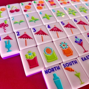 May include: A close-up of a set of white Mahjong tiles with colorful cocktail-themed illustrations. The tiles feature images of cherries, umbrellas, and cocktail glasses. The tiles are arranged on a red surface.