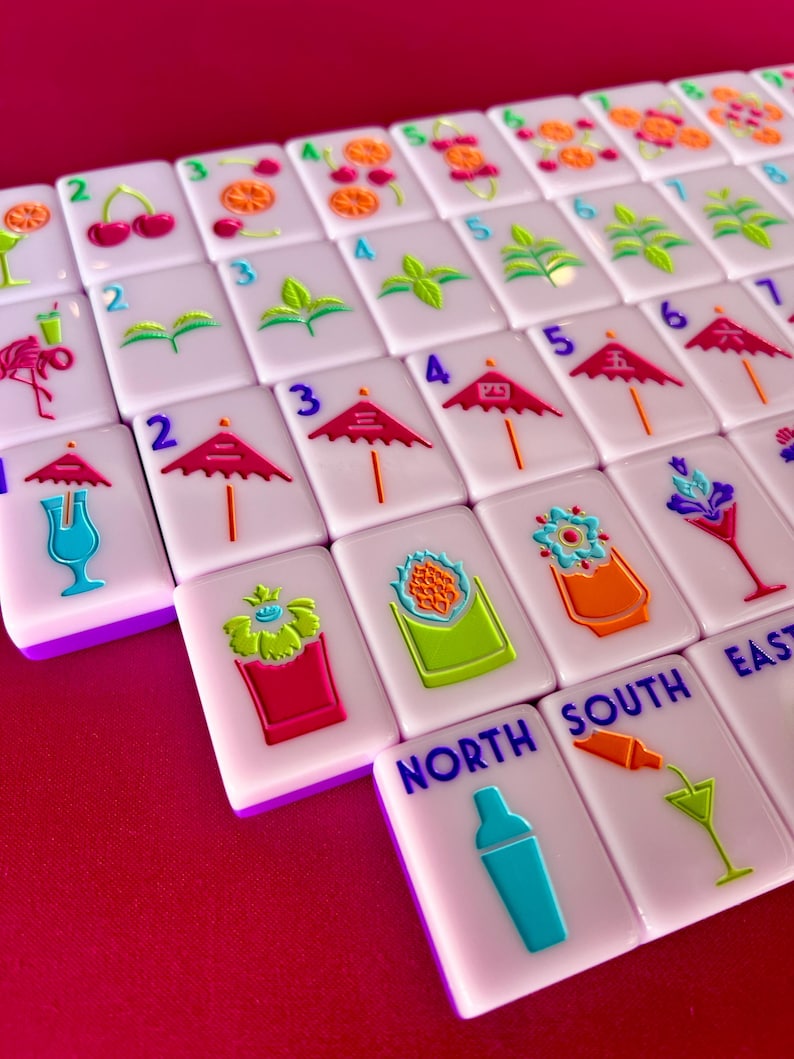 May include: A close-up of a set of white Mahjong tiles with colorful cocktail-themed illustrations. The tiles feature images of cherries, umbrellas, and cocktail glasses. The tiles are arranged on a red surface.