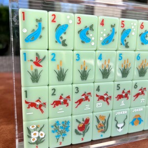 May include: A set of light green Mahjong tiles with various illustrations and numbers. The tiles feature images of fish, birds, reeds, horses, and flowers. Some tiles have the word "JOKER" on them.