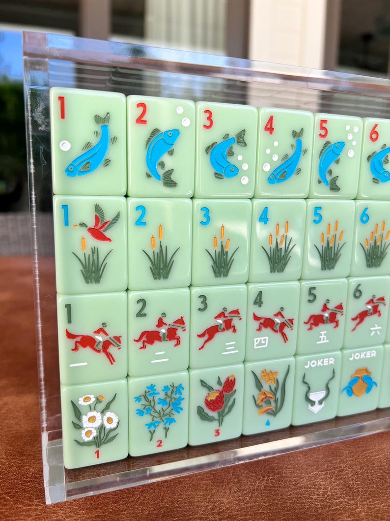 May include: A set of light green Mahjong tiles with various illustrations and numbers. The tiles feature images of fish, birds, reeds, horses, and flowers. Some tiles have the word "JOKER" on them.