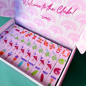 May include: A pink Mahjong set with tiles featuring cocktail and fruit illustrations, numbers, and directional markers. The box interior and lid display a pink fan pattern and the text "Welcome to the Club!" in cursive.