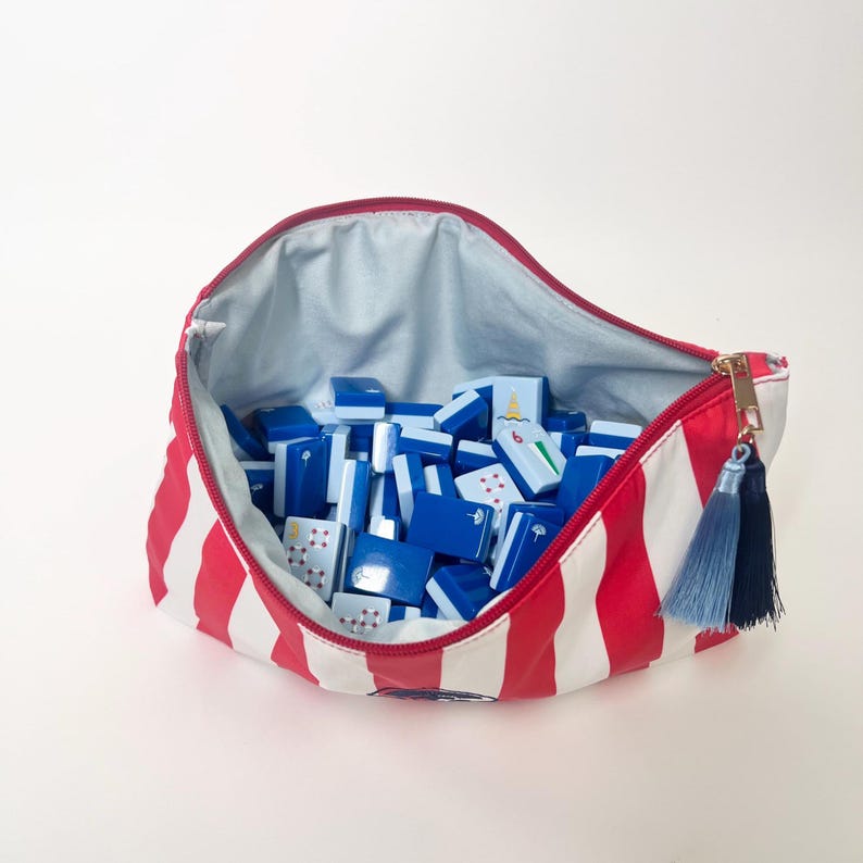 May include: A red and white striped zippered pouch filled with blue and white game tiles. The pouch has a gold zipper and a blue tassel. The tiles have various symbols and numbers. The pouch is open, revealing a light blue interior.
