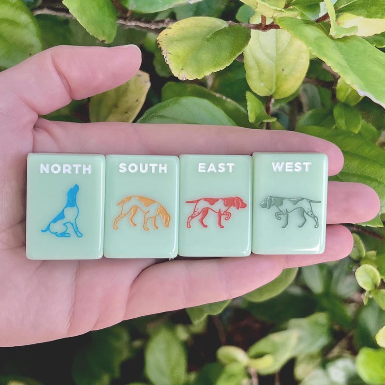 May include: Four light green rectangular tiles with the words NORTH, SOUTH, EAST, and WEST. Each tile features a dog illustration in blue, orange, red, and gray. The tiles are held in a hand.