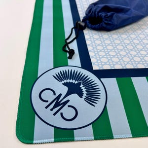May include: A blue, green, and white striped mat with a matching blue carrying bag. The mat features a circular logo with the letters "CMO" and a bird design. The mat is rolled up and secured with a black drawstring.