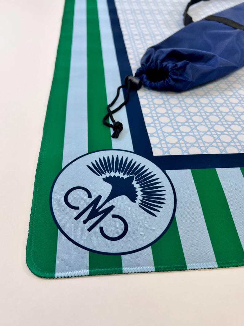 May include: A blue, green, and white striped mat with a matching blue carrying bag. The mat features a circular logo with the letters "CMO" and a bird design. The mat is rolled up and secured with a black drawstring.