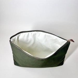 May include: An open, olive green zippered pouch with a light green and white gingham patterned interior. The zipper is black with a brown leather pull tab. The pouch is empty and sits against a white background.