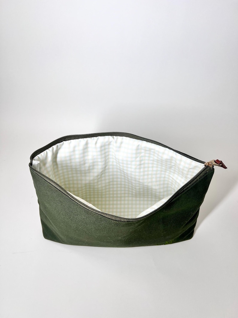 May include: An open, olive green zippered pouch with a light green and white gingham patterned interior. The zipper is black with a brown leather pull tab. The pouch is empty and sits against a white background.