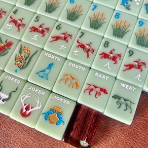 May include: A collection of light green Mahjong tiles with various embossed designs. The tiles feature numbers, Chinese characters, and images of fish, reeds, riders on horseback, and dogs. Some tiles are labeled "JOKER", "NORTH", "SOUTH", "EAST", and "WEST".