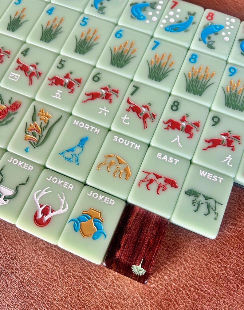 May include: A collection of light green Mahjong tiles with various embossed designs. The tiles feature numbers, Chinese characters, and images of fish, reeds, riders on horseback, and dogs. Some tiles are labeled "JOKER", "NORTH", "SOUTH", "EAST", and "WEST".