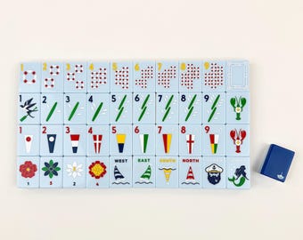 May include: A set of light blue rectangular mahjong tiles arranged in four rows, featuring various colorful illustrations including numbers, nautical flags, flowers, and maritime symbols. A single dark blue tile sits separately to the right.