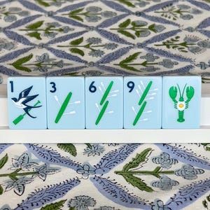 May include: Five light blue Mahjong tiles with white and green symbols and numbers. The tiles feature a bird, green stalks, and a green lobster. The tiles are arranged on a white surface, with a patterned blue and green fabric in the background.