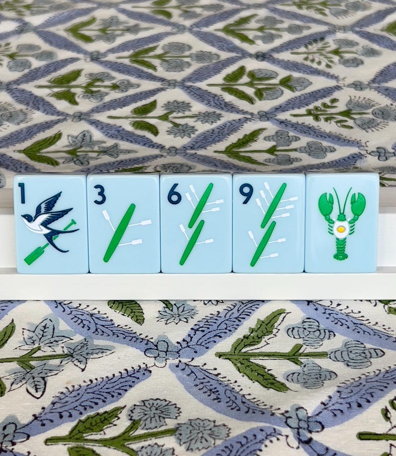 May include: Five light blue Mahjong tiles with white and green symbols and numbers. The tiles feature a bird, green stalks, and a green lobster. The tiles are arranged on a white surface, with a patterned blue and green fabric in the background.