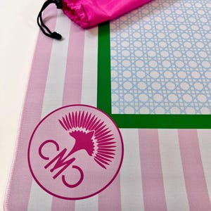 May include: A pink and white striped yoga mat with a green border and a light blue lattice design. A matching pink carrying bag is included. The mat features a circular logo with the letters "CM" and a stylized floral design.