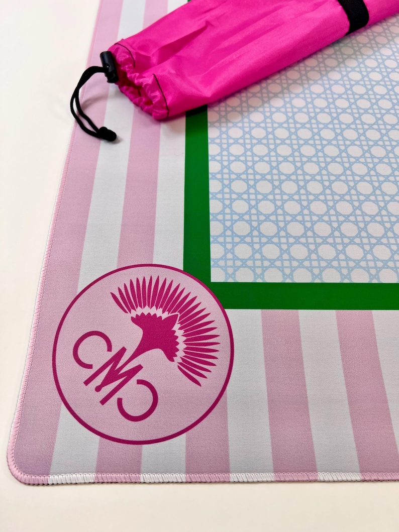 May include: A pink and white striped yoga mat with a green border and a light blue lattice design. A matching pink carrying bag is included. The mat features a circular logo with the letters "CM" and a stylized floral design.