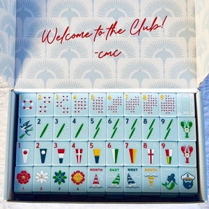 May include: A light blue Mahjong set with red and gold details, displayed in an open box. The tiles feature various symbols, numbers, and directional markers. The box lid has the text "Welcome to the Club!" in red.