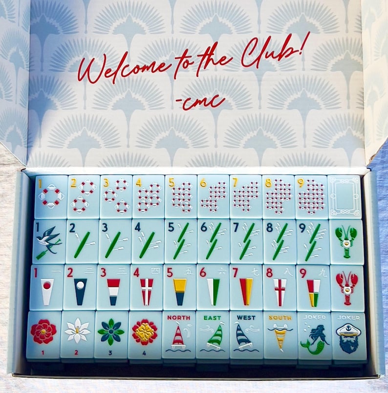 May include: A light blue Mahjong set with red and gold details, displayed in an open box. The tiles feature various symbols, numbers, and directional markers. The box lid has the text "Welcome to the Club!" in red.