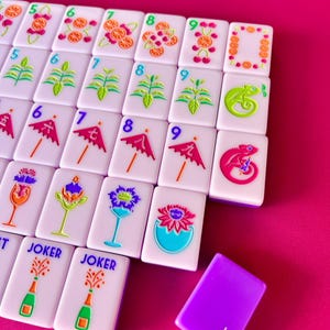 May include: A collection of white Mahjong tiles with colorful illustrations of fruits, plants, chameleons, and umbrellas. The tiles are arranged on a pink surface, with a purple tile in the foreground. Some tiles have the word "JOKER".