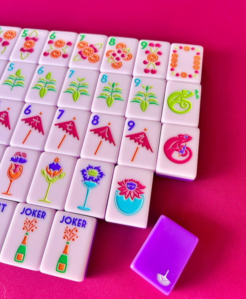 May include: A collection of white Mahjong tiles with colorful illustrations of fruits, plants, chameleons, and umbrellas. The tiles are arranged on a pink surface, with a purple tile in the foreground. Some tiles have the word "JOKER".