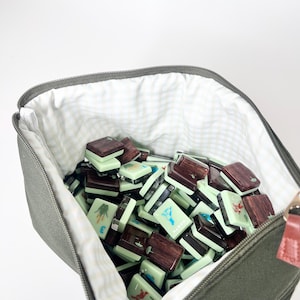 May include: A canvas bag in olive green, filled with a collection of mahjong tiles. The tiles are a mix of light green and dark brown, with various symbols and numbers. The bag has a gingham lining and a zipper closure.
