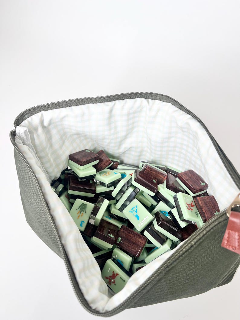 May include: A canvas bag in olive green, filled with a collection of mahjong tiles. The tiles are a mix of light green and dark brown, with various symbols and numbers. The bag has a gingham lining and a zipper closure.