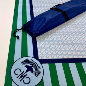 May include: A blue and green striped mat with a light blue cane weave pattern in the center. A blue carrying bag with a black strap is on top of the mat. A circular logo with the letters "CMO" and a bird graphic is in the corner.