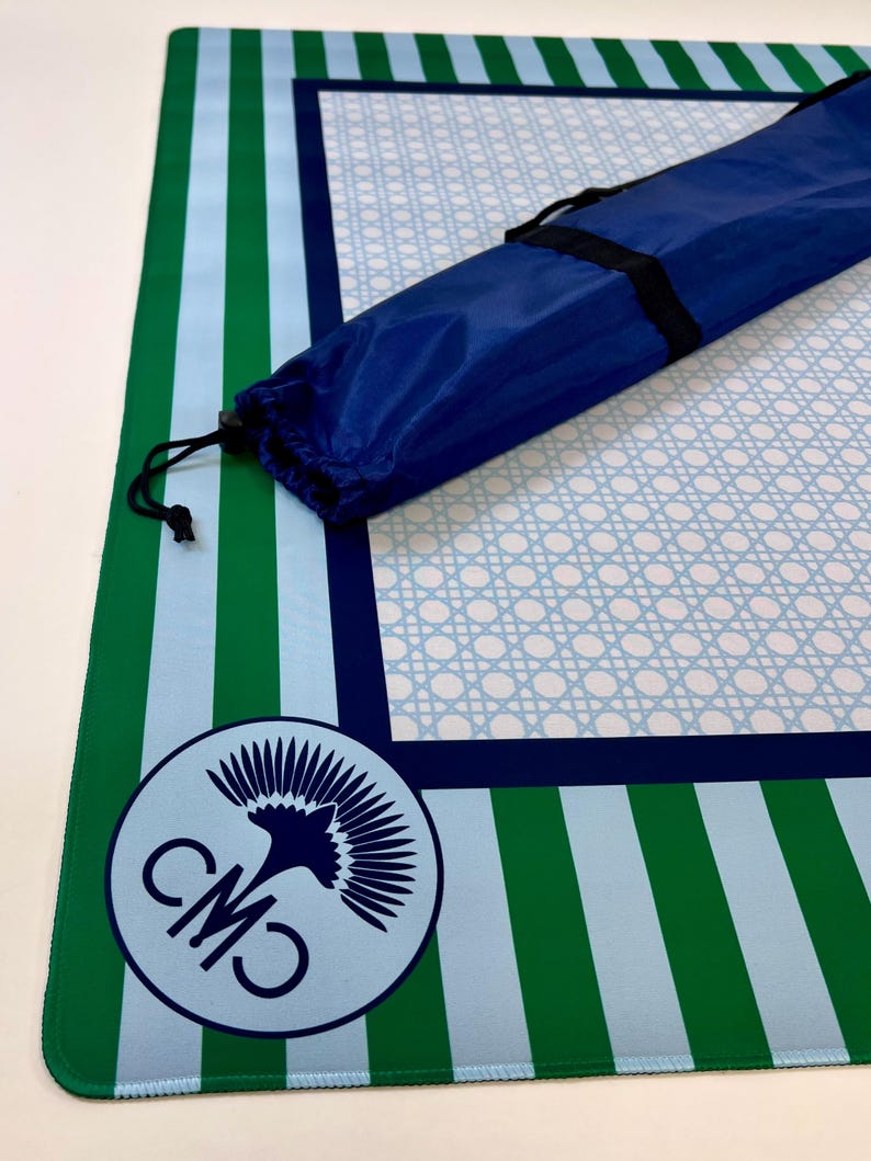 May include: A blue and green striped mat with a light blue cane weave pattern in the center. A blue carrying bag with a black strap is on top of the mat. A circular logo with the letters "CMO" and a bird graphic is in the corner.