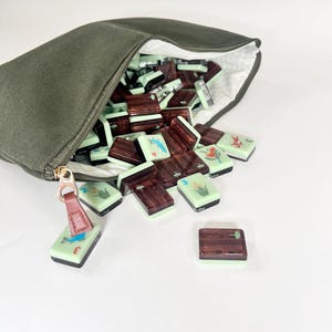 May include: An olive green canvas bag with a gold zipper, open to reveal a collection of mahjong tiles. The tiles are a mix of light green and dark brown, some with illustrations of birds and plants. A few tiles are scattered outside the bag.