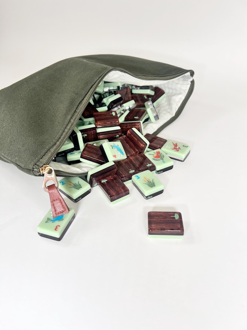 May include: An olive green canvas bag with a gold zipper, open to reveal a collection of mahjong tiles. The tiles are a mix of light green and dark brown, some with illustrations of birds and plants. A few tiles are scattered outside the bag.